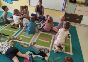 Zdjęcie dzieci i opiekunki siedzących na dywanie podczas przygotowań do zajęć pt. ,,POZNAJEMY POJAZDY''.