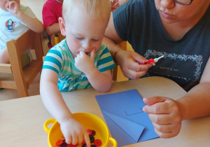 Zdjęcie zamyślonego Szymona z grupy Motylki podczas wybierania guziczków z żółtej miseczki do udekorowania swojej niebieskiej kartki na Dzień Taty.