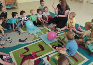 Opiekunka pokazuje dzieciom siedzącym w kółeczku na dywanie niebieski koralik w kształcie trójkąta.