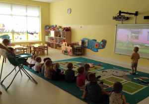 Zdjęcie dzieci siedzących na dywanie podczas oglądania bajki wyświetlanej na tablicy interaktywnej.