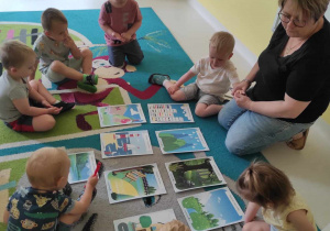 Zdjęcie opiekunki i dzieci siedzących na dywanie przy rozłożonych wydrukowanych obrazkach.