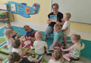 Zdjęcie opiekunki i dzieci siedzących na dywanie podczas czytania książeczki pt. ,,Kicia Kocia na wakacjach''.