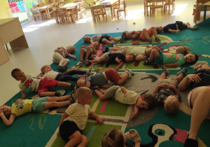 Zdjęcie dzieci i opiekunki z grupy Pszczółki leżących na dywanie, przygotowujących się do zajęć gimnastycznych w towarzystwie muzyki relaksacyjnej.