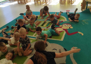 Dzieci razem z opiekunką ćwiczą na dywanie leżenie na brzuchu z rozłożonymi rękoma.