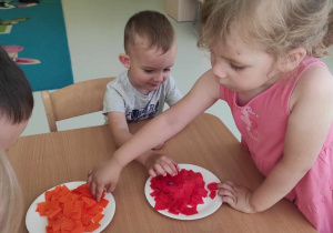 Alicja bierze z papierowego talerzyka pomarańczową bibułę a Nikodem bibułę w kolorze czerwonym.