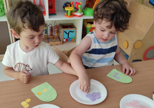 Wiktor i Bruno biorą z papierowego talerzyka fioletową aplikację w kształcie kwiatuszka.