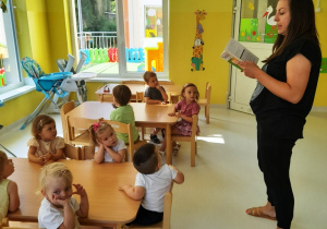 Opiekunka czyta dzieciom siedzącym przy stole wiersz.