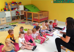 Dzieci siedząc na dywanie słuchają wiersza, który czyta opiekunka.