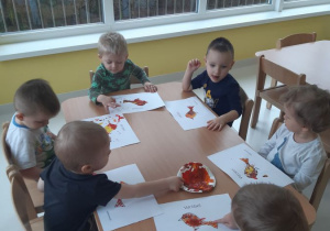 Dzieci siedzące przy stolikach wykonują pracę farbami.