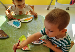 Dwoje dzieci przy stoliku malują pędzelkami i farbami elementy papierowej papugi.