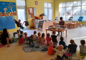 Zdjęcie opiekunki prowadzącej zajęcia i dzieci siedzących na dywanie.