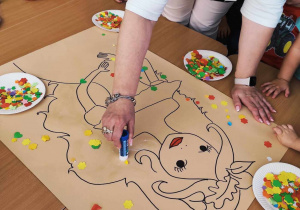 Jedna z opiekunek smaruje klejem miejsca, na których dzieci mają naklejać kolorowe aplikacje z papieru.