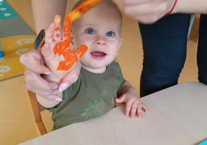 Opiekunka maluje pędzelkiem dłoń Nikodema pomarańczową farbą.