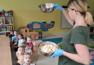 Opiekunka prezentuje dzieciom miseczki, w których znajdują się pokrojone w plasterki owoce.