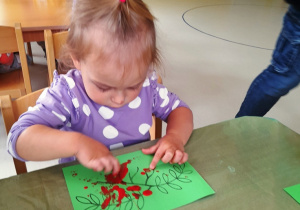 Ula maluje paluszkiem umoczonym w czerwonej farbie na zielonej kartce.