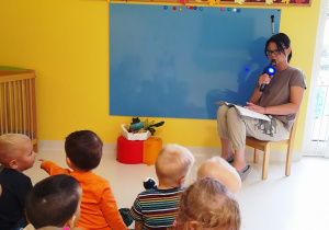 Opiekunka czyta wiersz dzieciom siedzącym na dywanie.