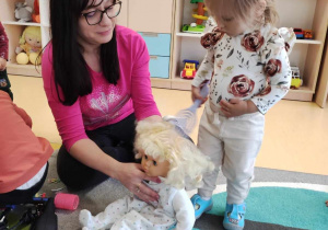 Opiekunka przytrzymuje Marcelinie lalkę, podczas gdy dziewczynka czesze jej włosy.