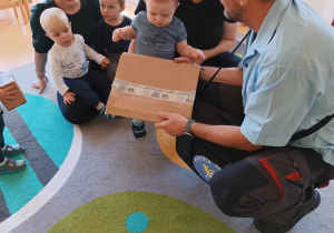Pan listonosz pokazuje dzieciom swoją przesyłkę w postaci tekturowej paczuszki.