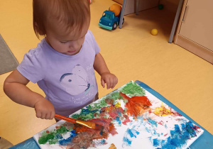 Laura maluje na płótnie krajobraz za pomocą pędzelka i kolorowych farb.