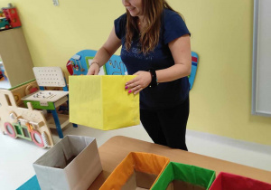 Zdjęcie opiekunki pokazującej dzieciom żółte tekturowe pudełko.