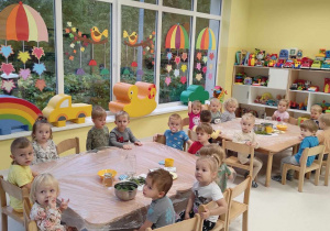 Zdjęcie dzieci siedzących przy stoliczku i przygotowujących się do zajęć kuchennych pt. ,,KISIMY OGÓRKI I MARYNUJEMY PAPRYKĘ''.
