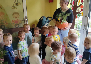 Zdjęcie opiekunki i dzieci stojących na korytarzu przed drzwiami wejściowymi do żłobka z wykonanymi jesiennymi dyniowymi wazonami.