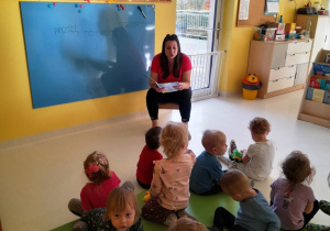 Opiekunka czyta dzieciom siedzącym na dywanie wiersz.