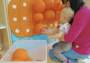 Amelka siedząca na kolanach opiekunki, przykleja z jej pomocą pomarańczowy balonik.