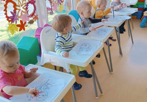Maluszki siedzące w dziecięcych foteliczkach malują kolorowymi kredkami swoje kolorowanki.