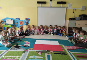 Pamiątkowe zdjęcie dzieci z grupy Pszczółki siedzących na dywanie z przyczepionymi na koszulkach kotylionami i ułożoną z białych i czerwonych kartek papieru flagą Polski.