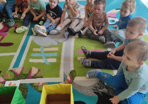 Zdjęcie dzieci siedzących na dywanie słuchających tłumaczącej opiekunki, na czym będą polegać przeprowadzane zajęcia.