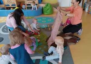Opiekunka pokazuje dzieciom siedzącym na dywanie rozłożoną chustę wykonaną z włóczki.
