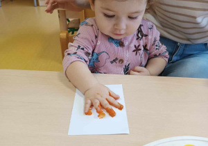 Zdjęcie Mai podczas odciskania swojej dłoni pomalowanej pomarańczową farbą na kartce.