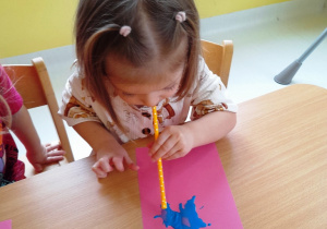 Mała dziewczynka próbuje za pomocą słomki stworzyć kałużę z niebieskiej farby.
