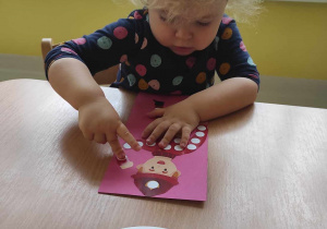 Skupiona Laura przykleja na Strażaka Sama przyklejonego na różowej kartce kolorową kuleczkę z plasteliny.