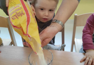 Szymon z pomocą opiekunki wsypuje do szklanki mąkę.