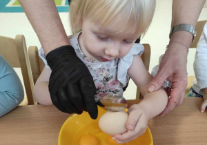 Opiekunka pomaga Aleksandrze rozbić łyżeczką jajko.