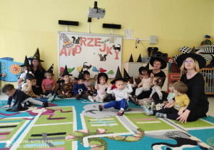Pamiątkowe zdjęcie dzieci i opiekunek z grupy Pszczółki siedzących na dywanie na tle udekorowanej tablicy interaktywnej z okazji Andrzejek z założonymi na głowach czarnymi kapeluszami Czarownic.