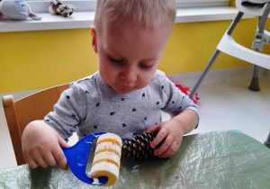Chłopczyk maluje szyszkę na biało za pomocą farby i wałeczka.