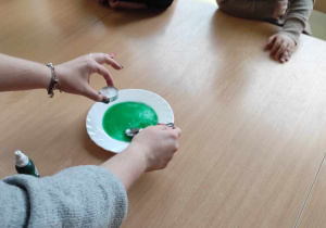 Opiekunka miesza za pomocą łyżeczki na talerzyku, wodę z dodatkiem zielonego barwnika.