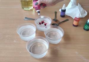 Opiekunka dodaje do plastikowych okrągłych miseczek z wodą po parę kropelek różnokolorowych barwników.