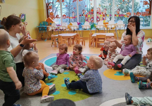 Zdjęcie dzieci i opiekunek z grupy Motylki siedzących na dywanie, wysyłających pozdrowień za pomocą wysyłanych buziaczków podczas zajęć pt. ,,DZIEŃ ŻYCZLIWOŚCI I POZDROWIEŃ''.