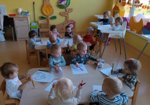 Zdjęcie dzieci siedzących przy stoliczku podczas kolorowania kolorowanek wylosowanymi przez siebie kredeczkami.