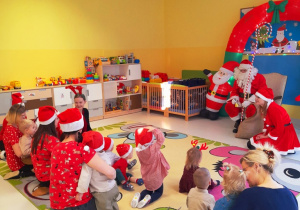 Dzieci wraz opiekunkami siedząc na dywanie przyglądają się Mikołajowi.