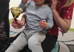 Zdjęcie Jakuba z założonymi świątecznymi okularami, siedząc na kolanach jednej z opiekunek trzyma złoty dzwoneczek.