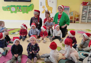 Dzieci oraz opiekunki siedzące na dywanie oczekują przyjścia Świętego Mikołaja.