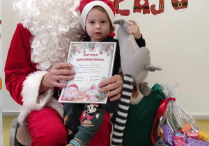 Szymon siedzący na kolanach Mikołaja ze swoją ulubioną zabawką i dyplomem pozuje do pamiątkowego zdjęcia.