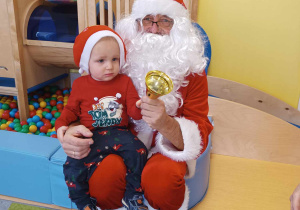 Pamiątkowe zdjęcie Tobiasza siedzącego na kolanach u Świętego Mikołaja ze złotym dzwoneczkiem trzymanym w dłoni.