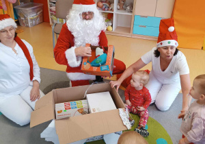 Mikołaj pokazuje maluszkom przyniesione przez siebie prezenty.