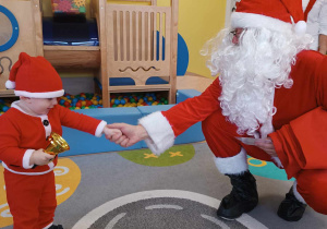 Święty Mikołaj podaje rączkę małemu Mikołajowi, który trzyma w dłoni złoty dzwoneczek.